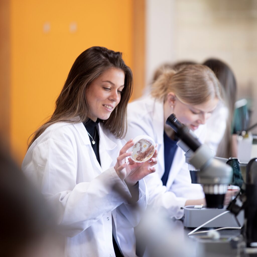 En elev fra Campus Odsherred står iført kittel foran et mikroskop. Elev kigger på en prøve i et naturvidenskabeligt fag.