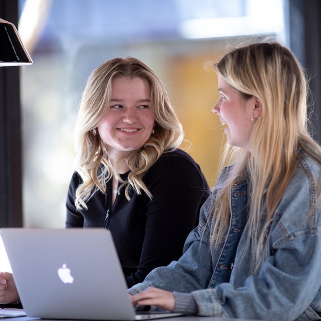 To elever fra Campus Odsherred kigger på hinanden og smiler. Den ene pige har en computer stående foran sig.