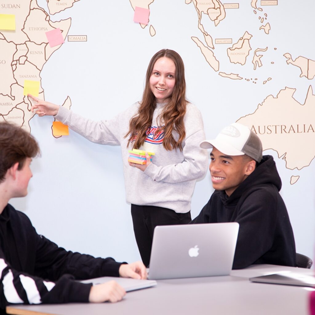 Tre elever fra Campus Odsherred sidder med en computer i klassen. Den ene elev står og peger på et land på verdenskortet, der hænger på væggen.