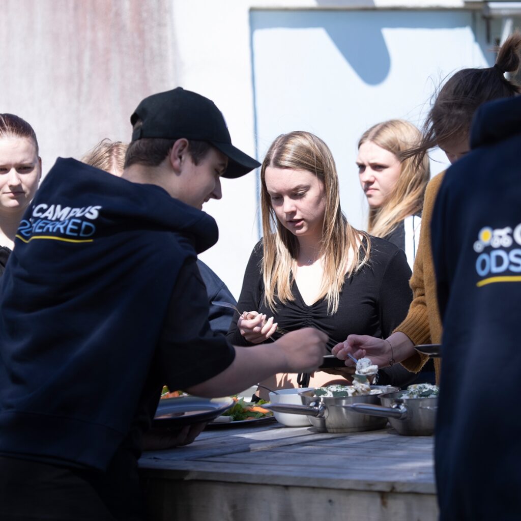 Elever fra Campus Odsherred står udenfor ved et bord og tager mad på deres tallerkener.