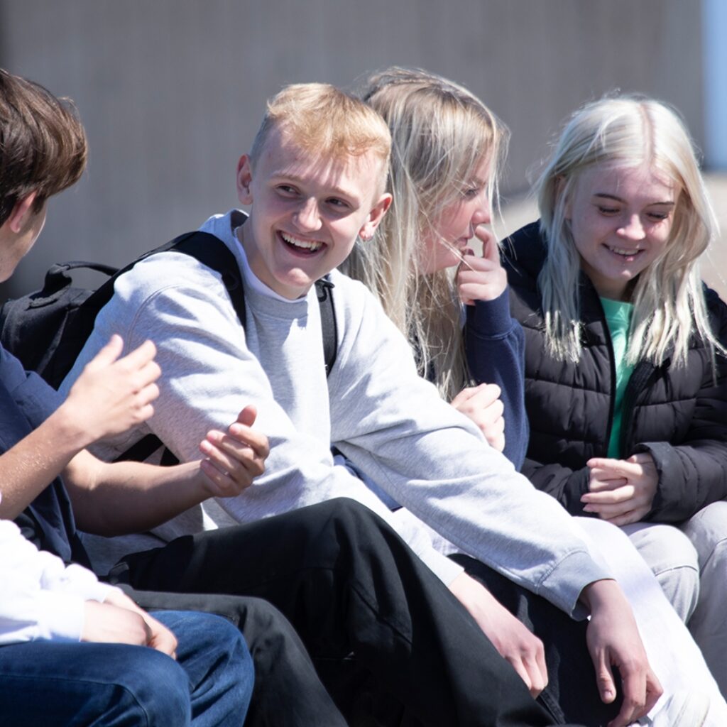 En gruppe elever fra Campus Odsherred sidder udenfor og snakker. Kameraret fokuserer på en dreng der ser glad ud.