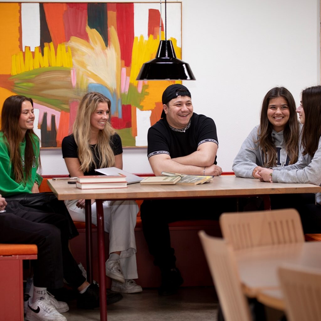 Fem elever fra Campus Odsherred sidder ved et bord i fællesrummet. De ser alle glade ud.